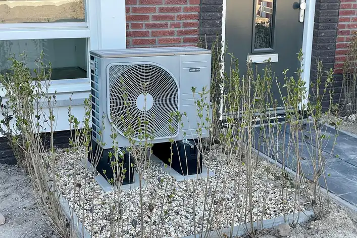 Unidad exterior de una bomba de calor junto a la pared de una casa, sobre un lecho de grava y rodeada de arbustos jóvenes