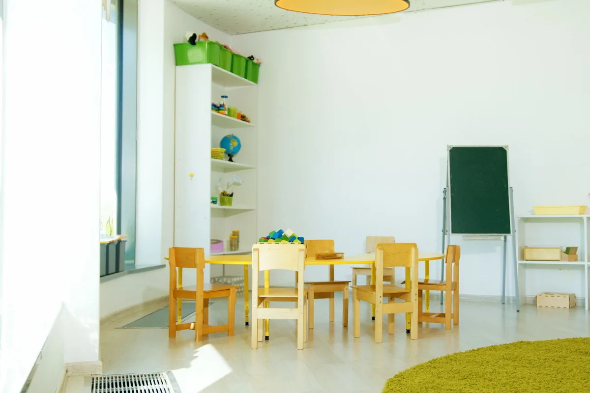 Aula infantil luminosa con una mesa amarilla, sillas de madera pequeñas, estanterías y una pizarra verde