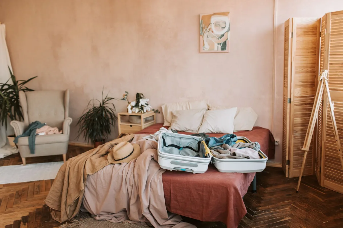 Dormitorio con la cama deshecha y una maleta abierta encima, junto a un sillón y un biombo de madera