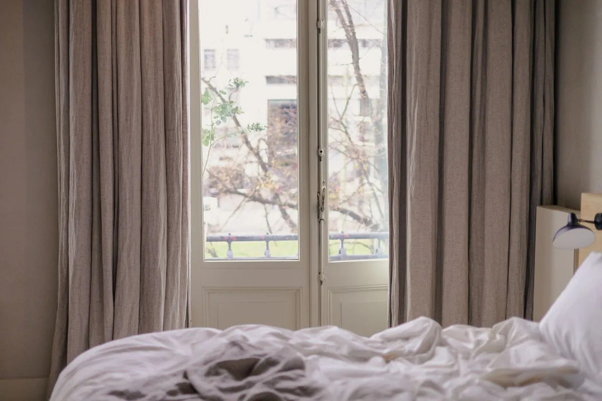 Dormitorio con la cama deshecha junto a una puerta de balcón con cortinas de lino