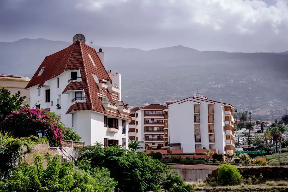Edificio blanco con tejado de teja y bloques de pisos con balcones de madera, con una montaña al fondo