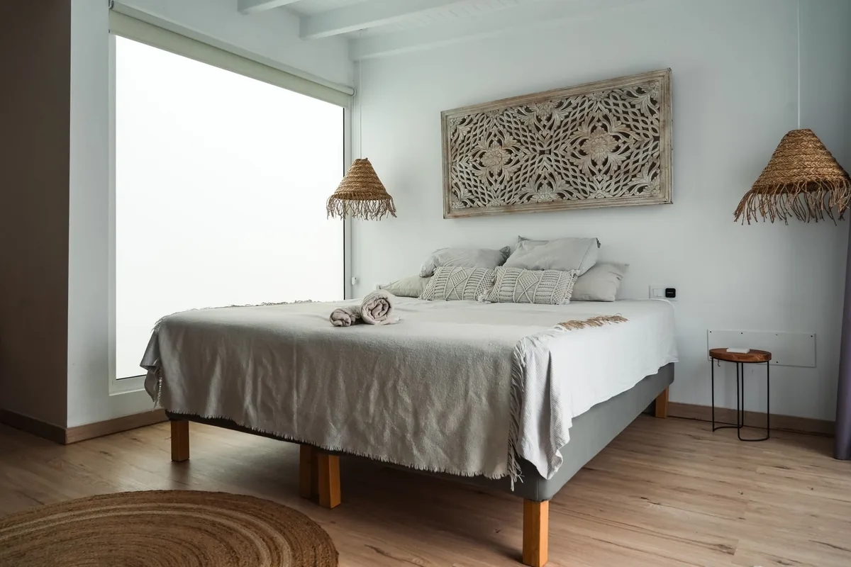 Dormitorio blanco y luminoso con cama doble, lámparas de rafia y suelo de madera clara