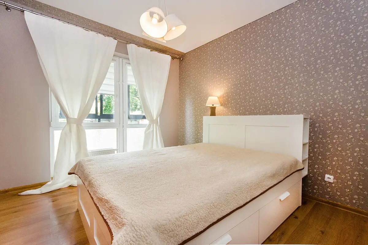 Dormitorio con cama de cabecero blanco, cortinas claras y una lámpara de mesa encendida