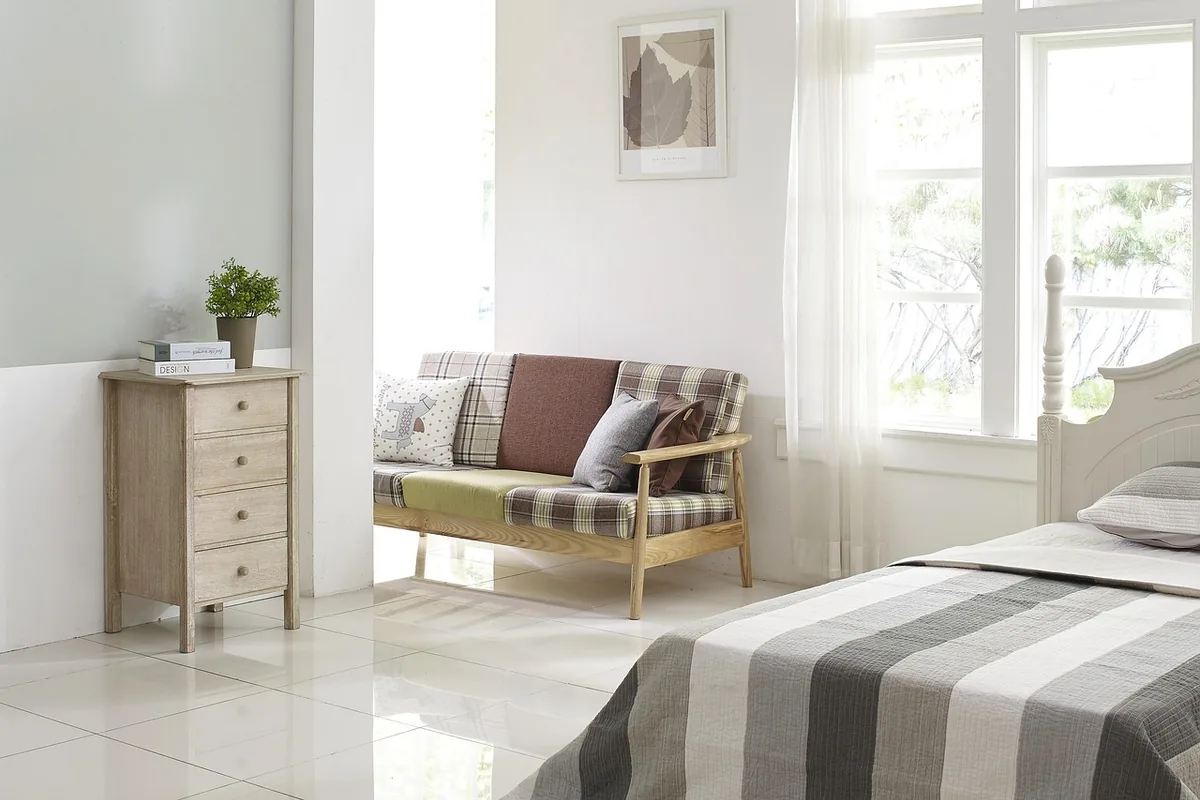 Habitación luminosa con una cama individual, un sofá de madera con cojines y una ventana con cortina blanca