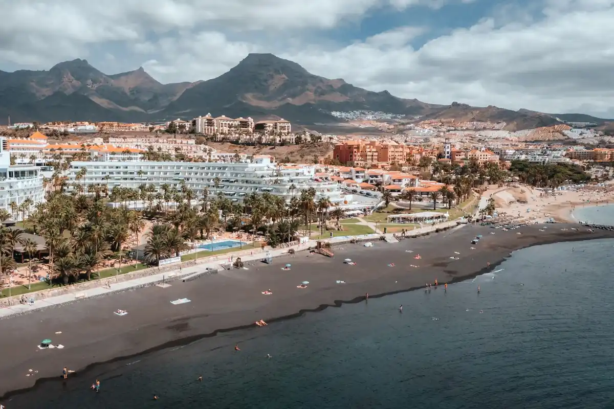 Vista aérea de la costa de Adeje con una playa de arena oscura, hoteles, palmeras y montañas al fondo