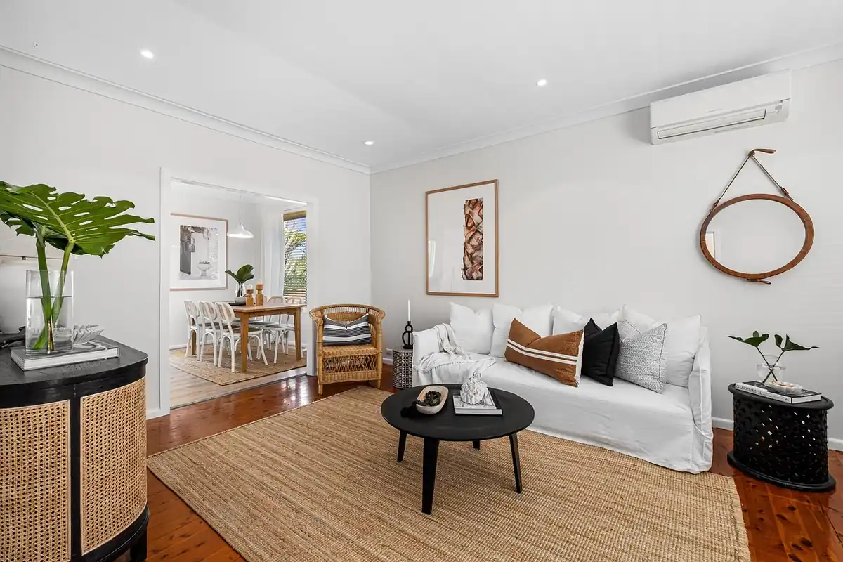 Salón luminoso con un sofá blanco y un split de pared sobre él, abierto a un comedor con mesa de madera