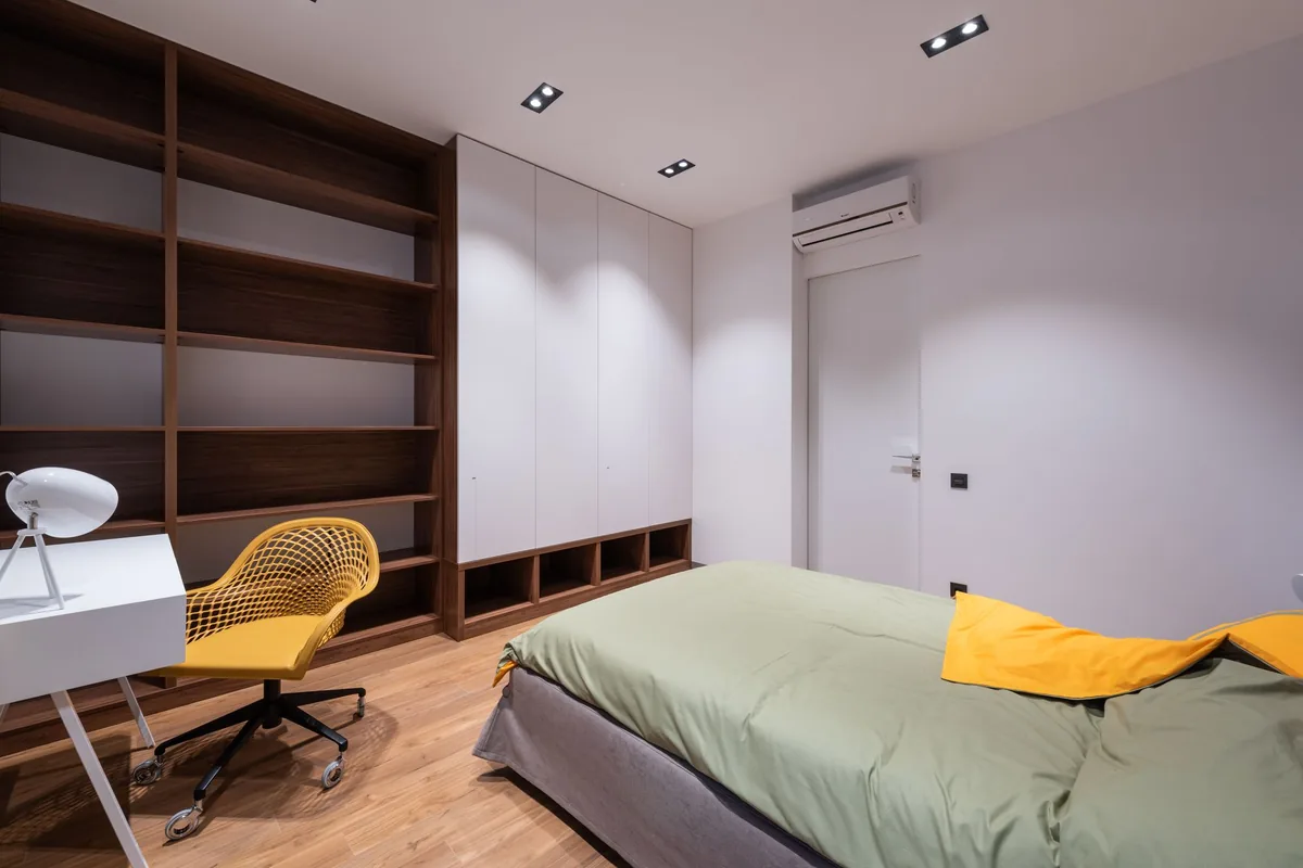 Dormitorio con una cama, una librería de madera, una silla amarilla y un split blanco en la pared