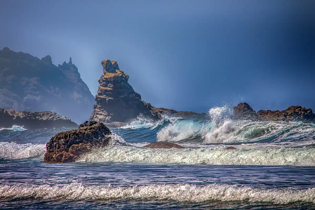 Olas rompiendo contra las rocas de una costa, con espuma blanca y un peñasco al fondo