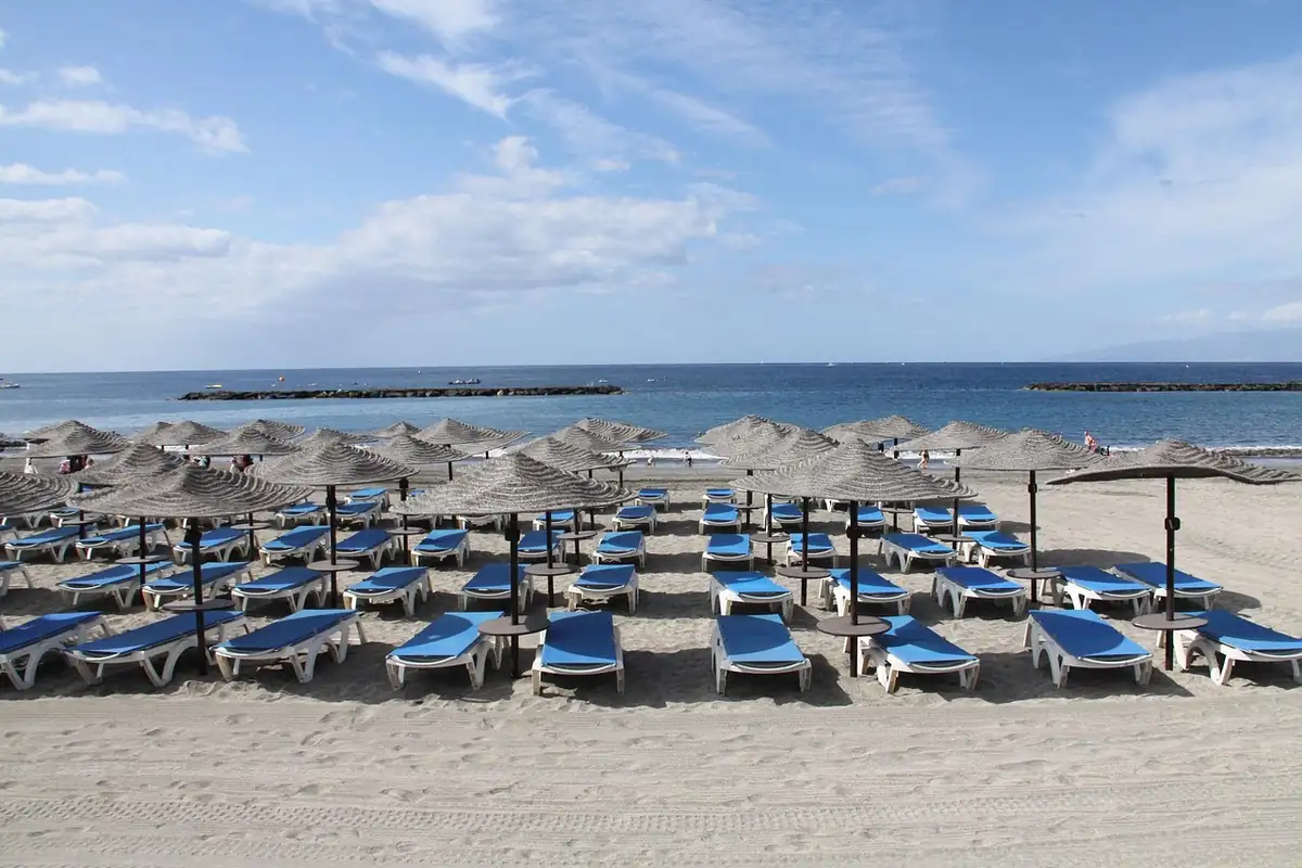 Playa de arena con sombrillas de paja y tumbonas azules vacías frente al mar bajo un cielo con nubes
