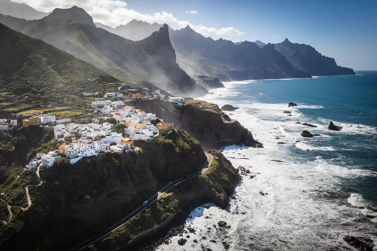 Vista aérea de un pueblo de casas blancas sobre un acantilado, entre montañas verdes y el mar, en las islas Canarias