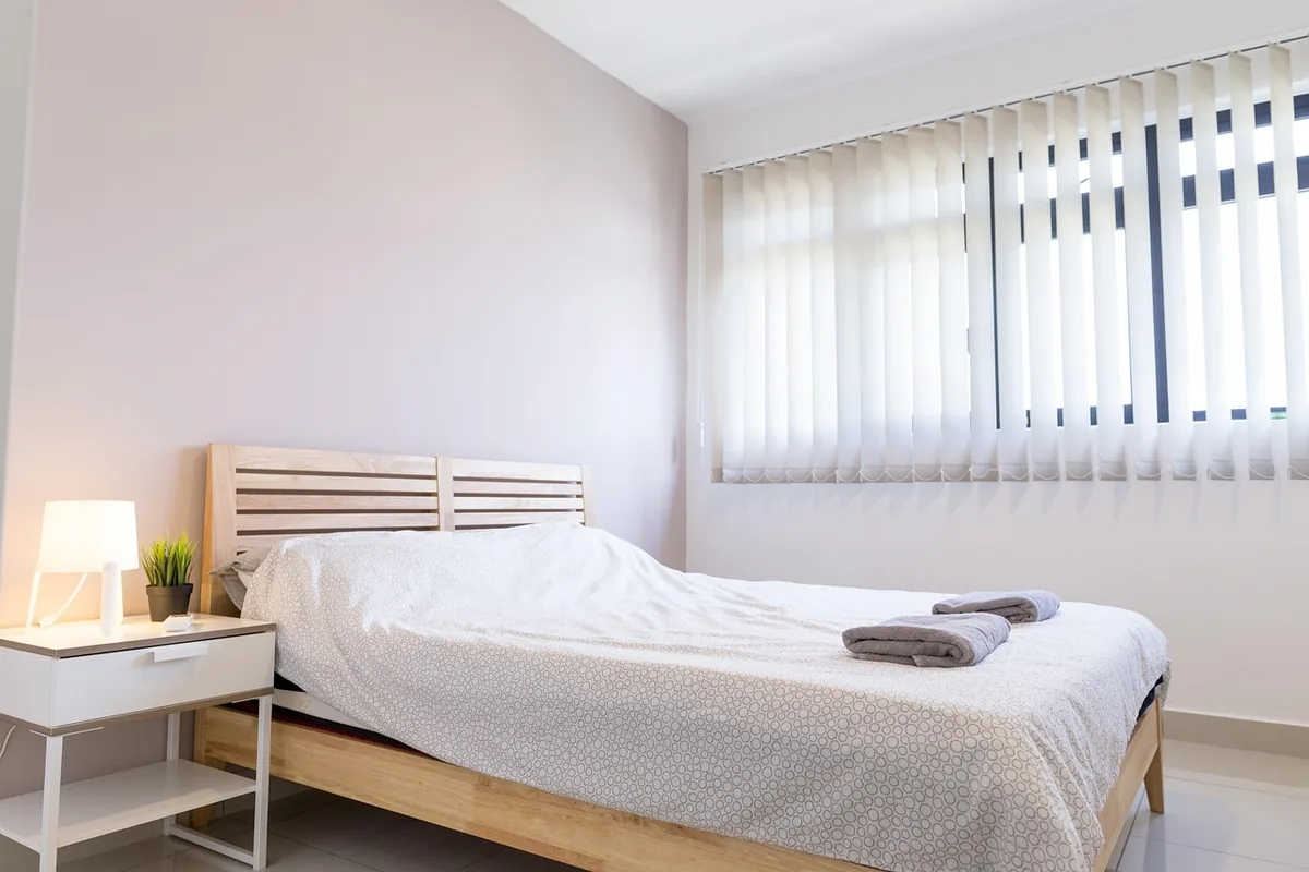 Dormitorio luminoso con una cama, una mesita de noche y una ventana con cortinas verticales