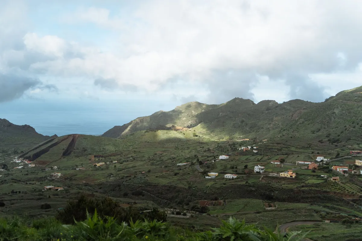 Ladera verde con casas dispersas, bancales y nubes bajas, con el mar al fondo