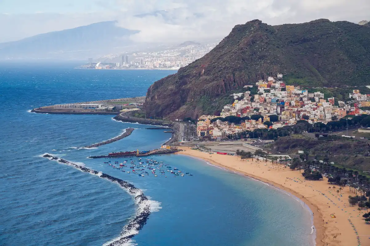 Vista aérea de una playa de arena con un pueblo de casas de colores al pie de una montaña, en la costa de Tenerife