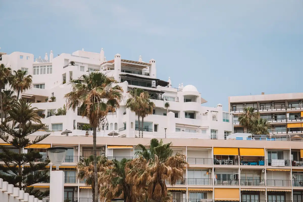 Complejo de apartamentos blancos entre palmeras, con terrazas y toldos amarillos bajo un cielo azul