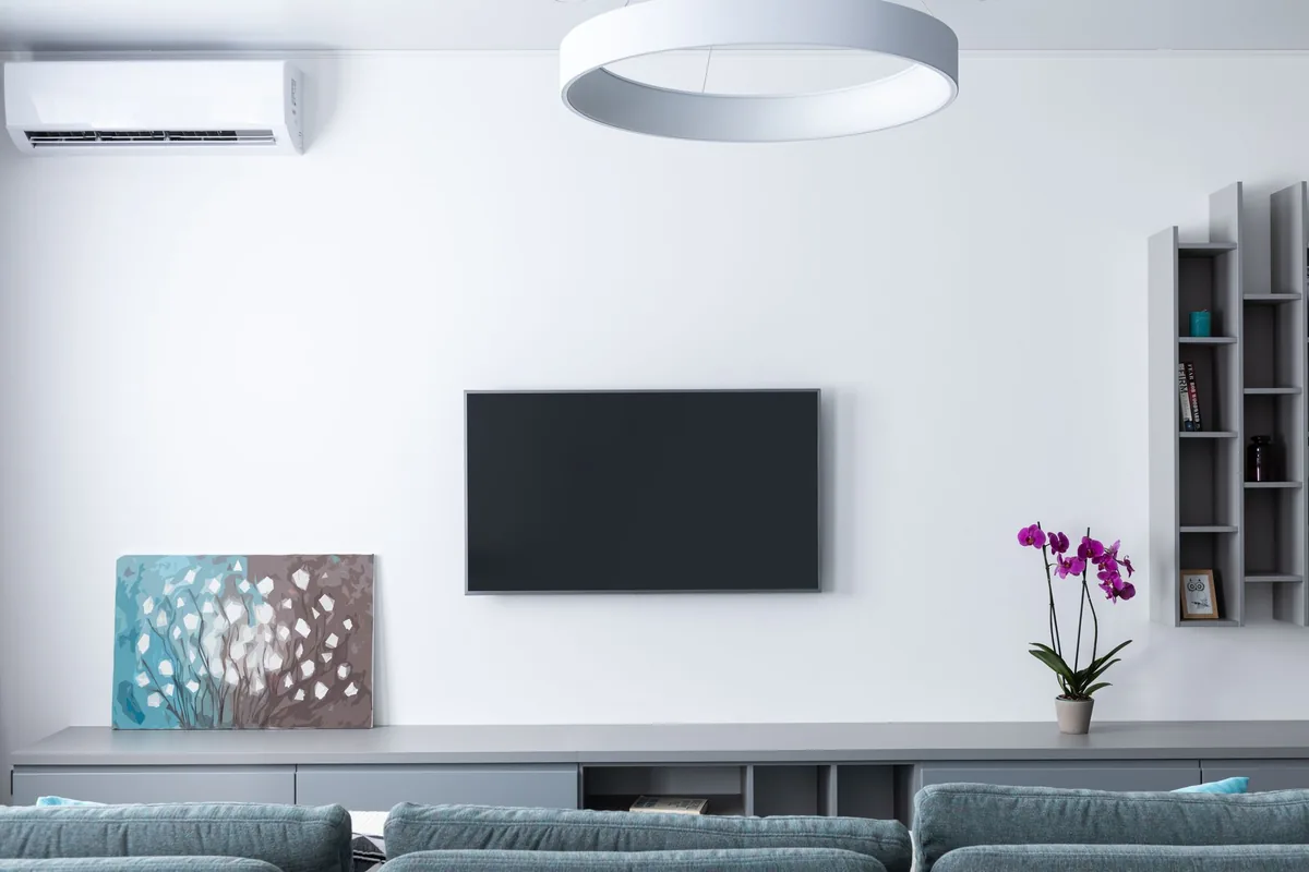 Salón luminoso con un split de pared en lo alto, un televisor apagado y un sofá gris
