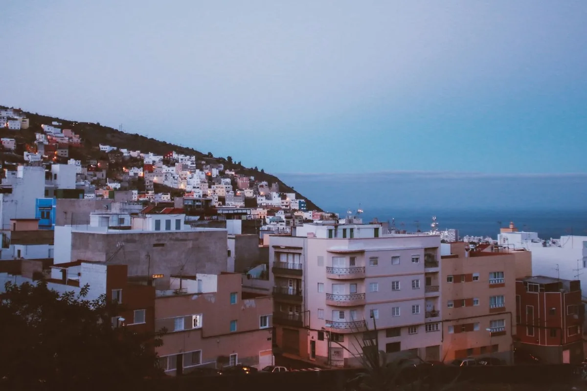 Pueblo en una ladera con un bloque de pisos, casas bajas y el mar al fondo al anochecer