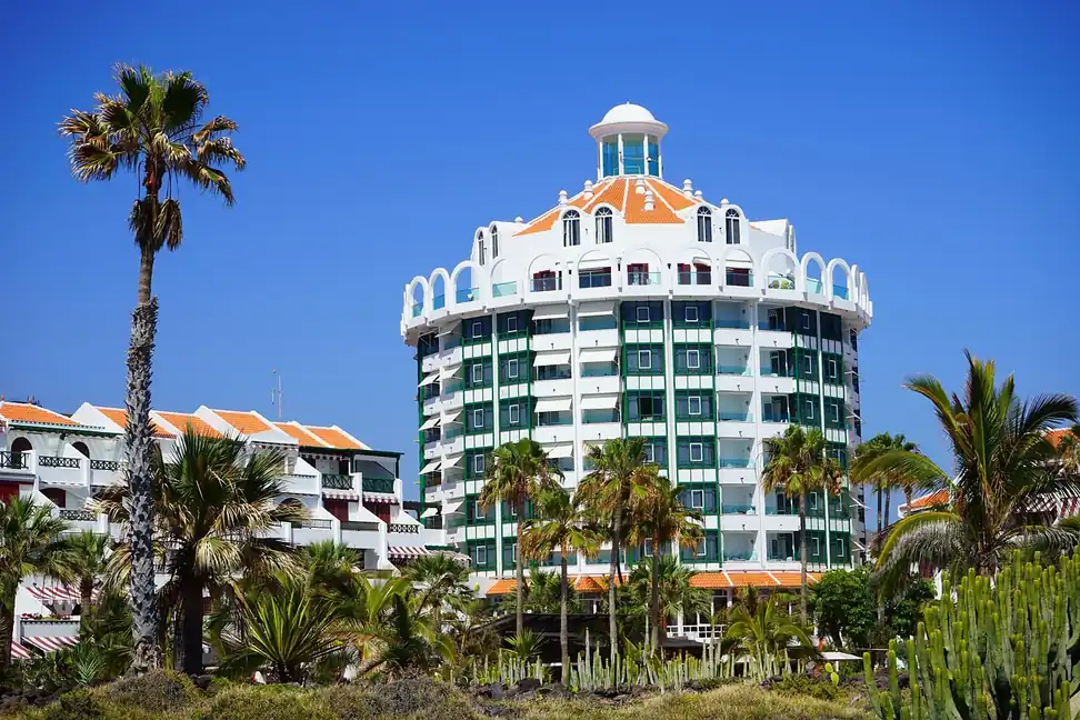 Edificio blanco de varias plantas con balcones, entre palmeras y cactus, bajo un cielo azul