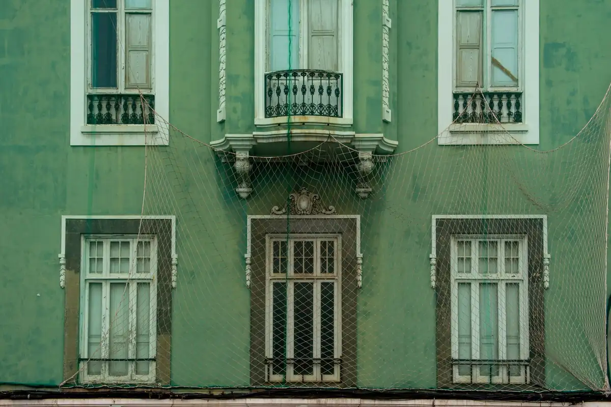 Fachada verde de un edificio antiguo con ventanas de madera y balcones de hierro, cubierta por una red