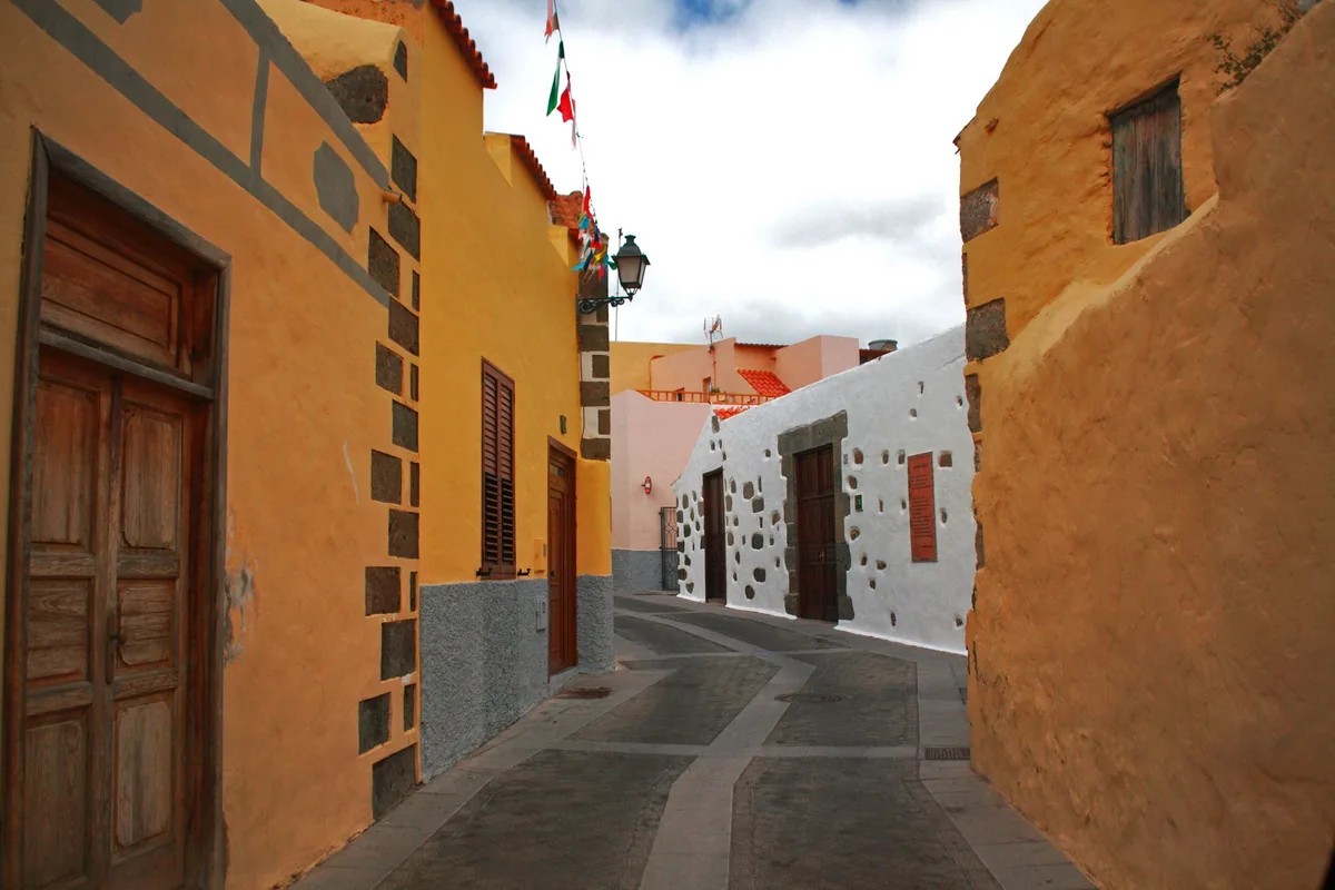 Calle estrecha de un pueblo con casas pintadas de amarillo y blanco, puertas de madera y banderines
