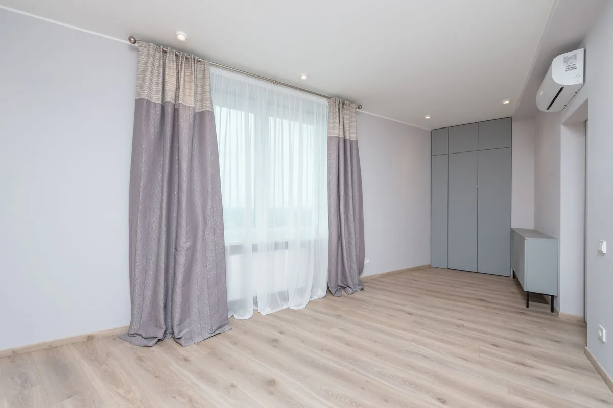 Habitación vacía con suelo de madera clara, cortinas grises y un split de pared junto al techo