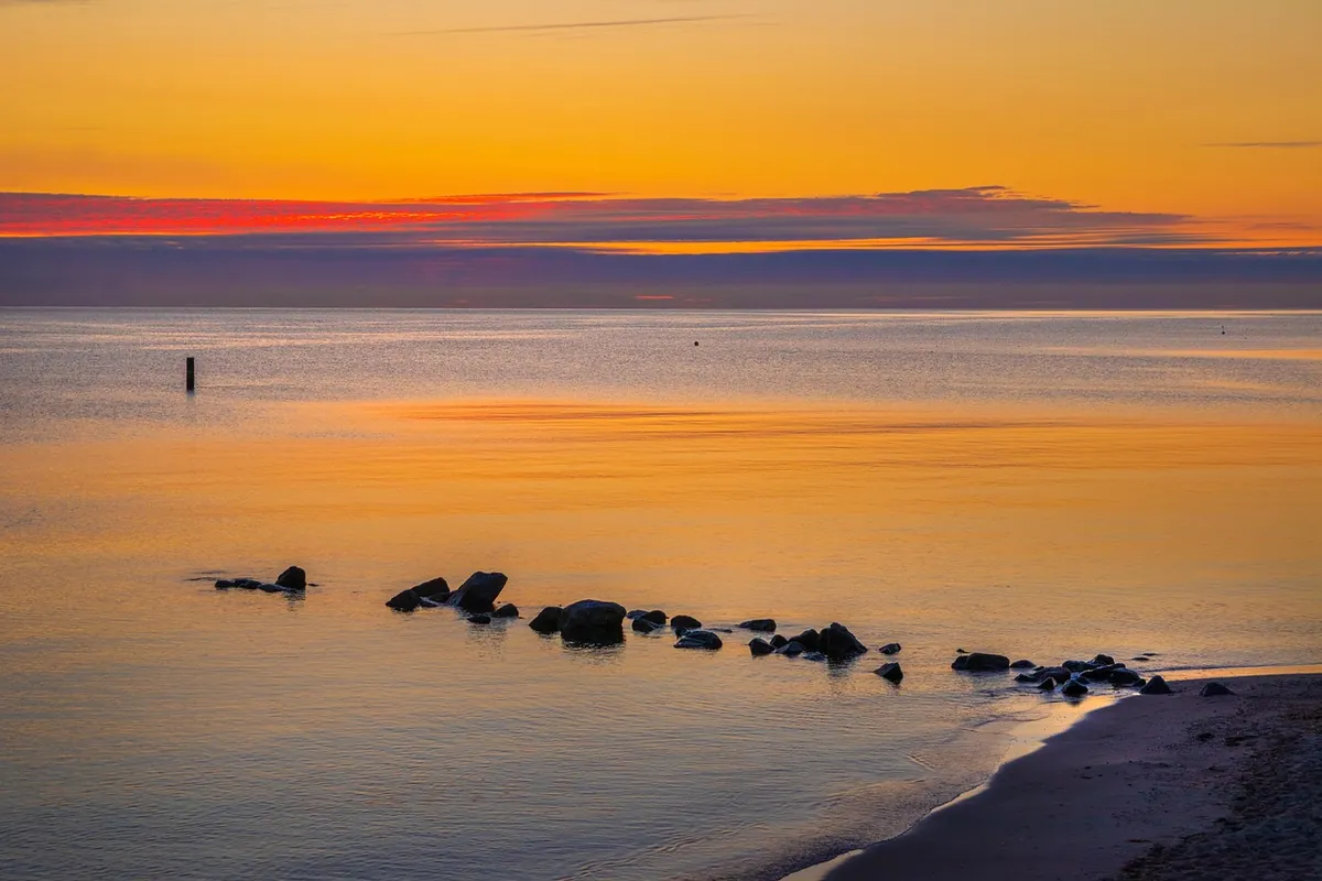Mar en calma hasta el horizonte bajo un cielo naranja y violeta, con unas rocas junto a la orilla